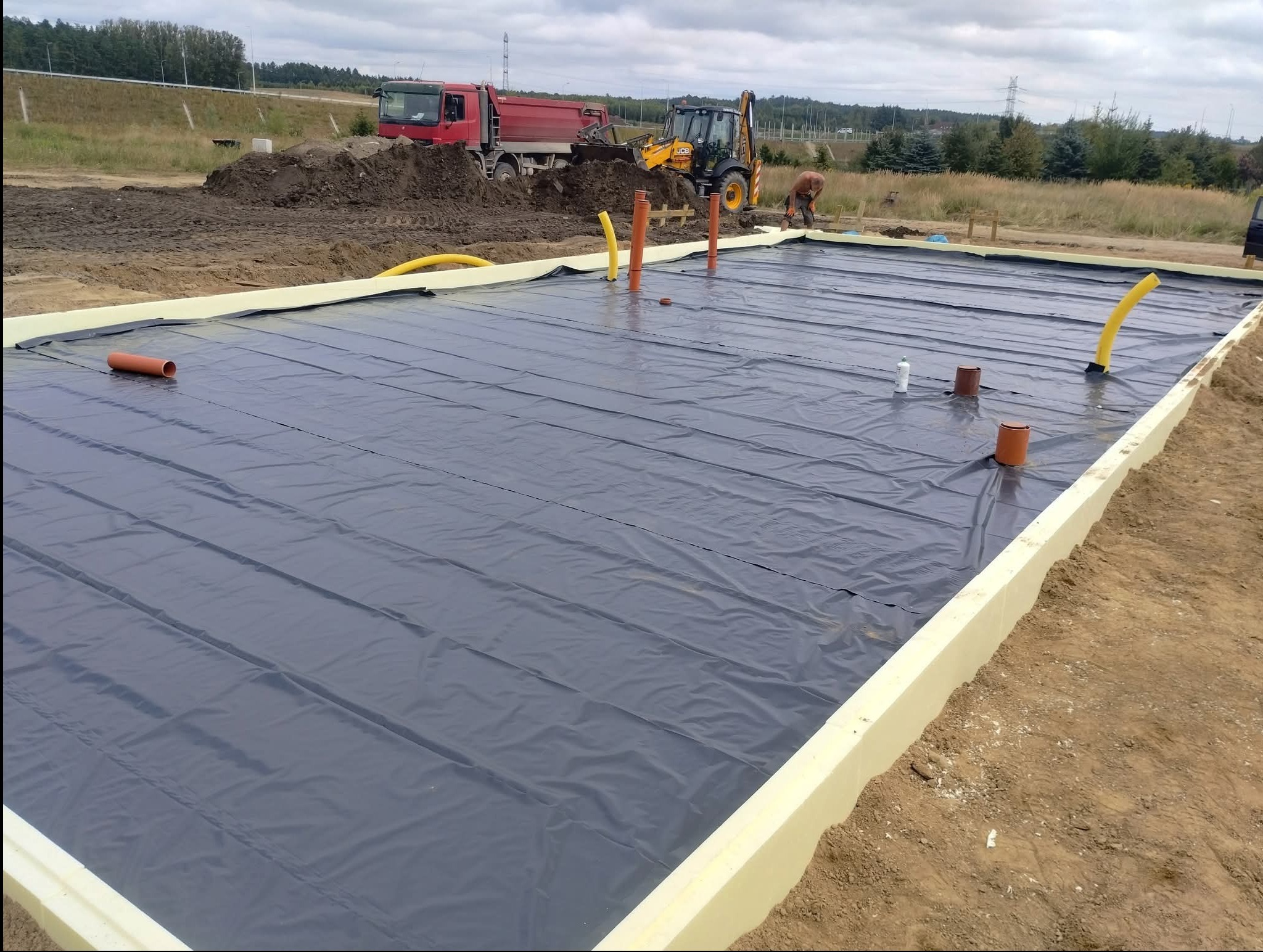 Przygotowany teren pod fundamenty z izolacją termiczną i folią hydroizolacyjną. W tle ciężarówka i koparka, widoczny pracownik.