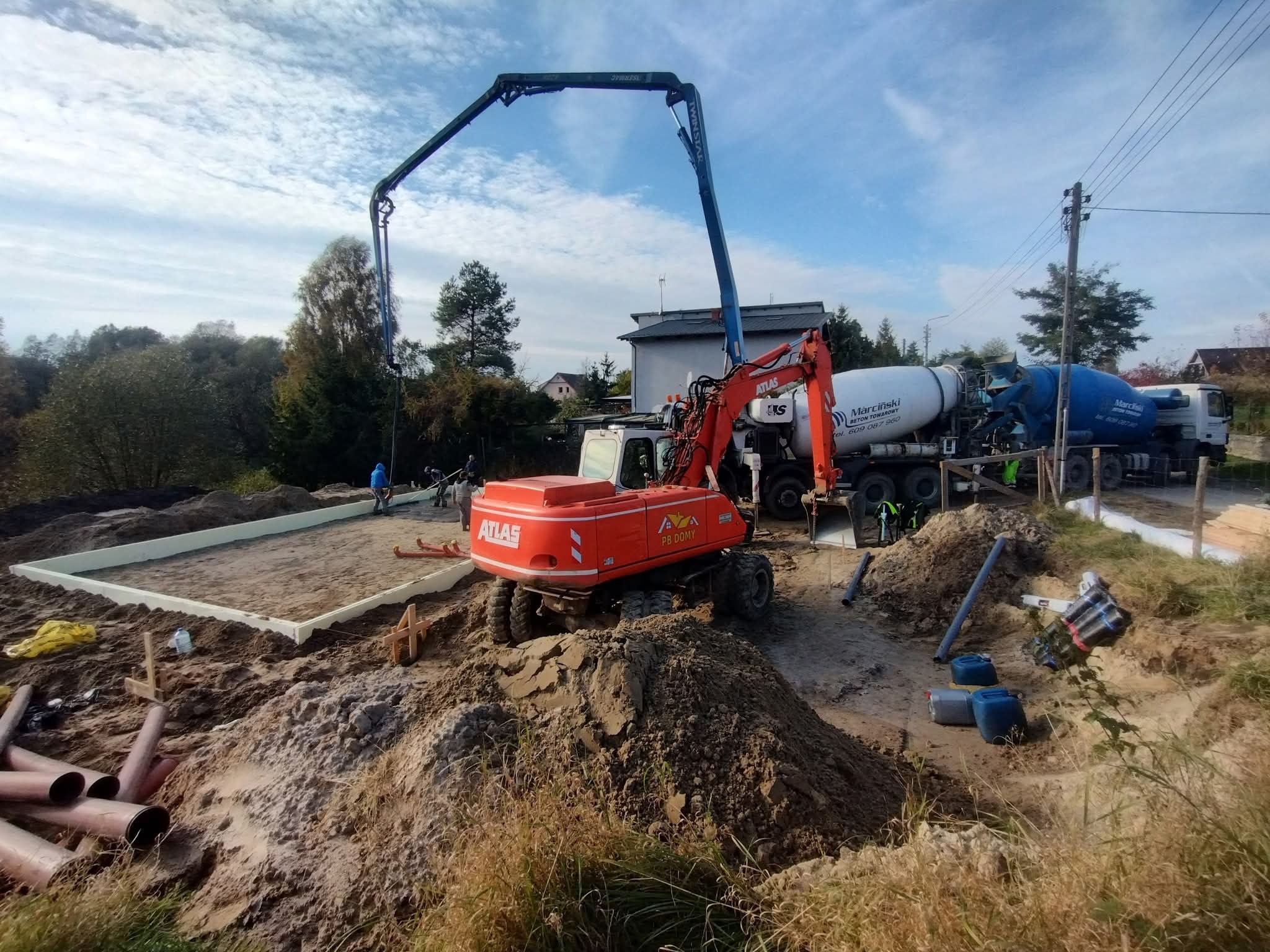 Wylewka fundamentów: pomarańczowa pompa do betonu, betoniarka i wykop z izolacją. Widoczne przygotowania do zalewania fundamentów pod budowę domu.