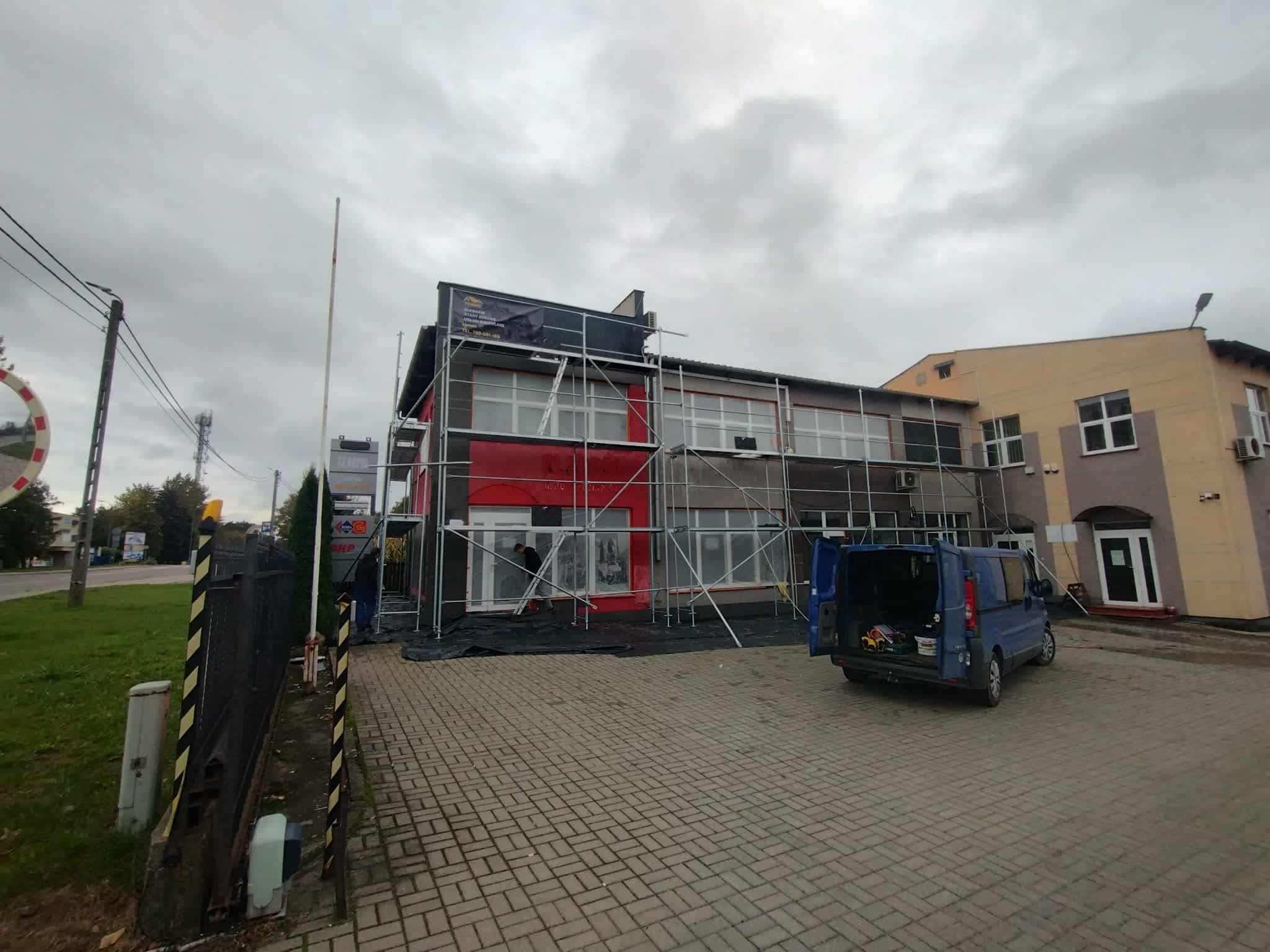 Budynek komercyjny w trakcie renowacji elewacji, otoczony rusztowaniami. Widoczny samochód dostawczy i fragment chodnika z kostki brukowej. Pochmurne niebo.