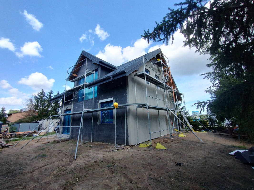 Dwukondygnacyjny dom w trakcie ocieplania elewacji, z widocznymi rusztowaniami i zabezpieczeniami okien folią. Częściowo otynkowana ściana, niebieskie niebo z chmurami w tle.