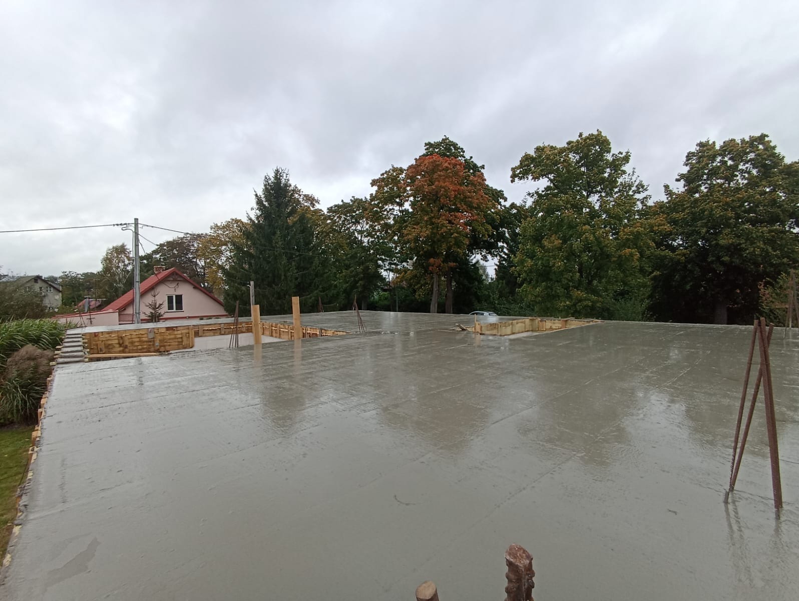 Świeżo wylana, mokra płyta fundamentowa z drewnianym szalunkiem w tle, widok na dom i drzewa pod pochmurnym niebem. Widoczne pręty zbrojeniowe.