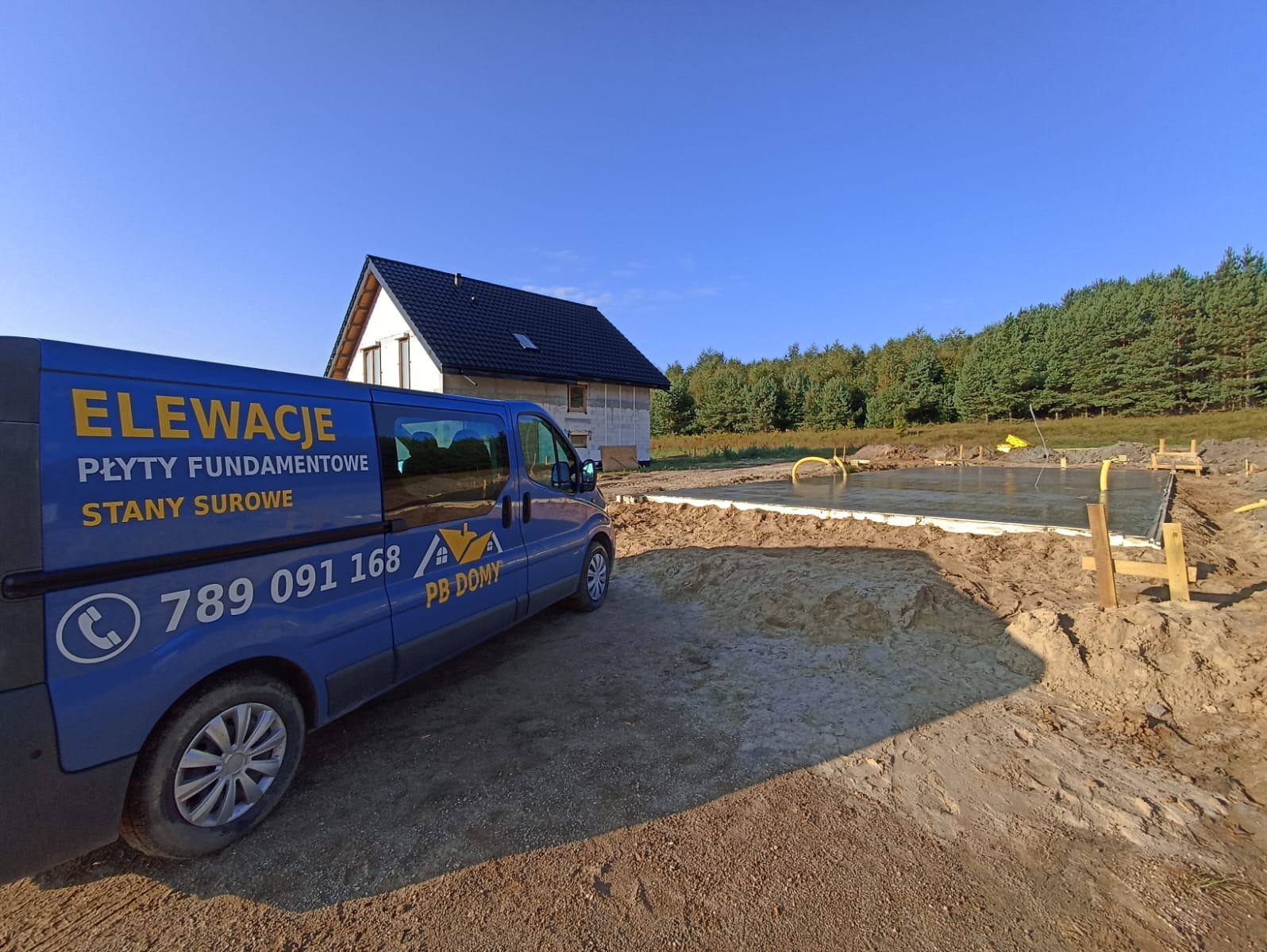 Wylana płyta fundamentowa pod budowę domu jednorodzinnego, widoczny firmowy bus z reklamą elewacji, stan surowy, w tle las i błękitne niebo.