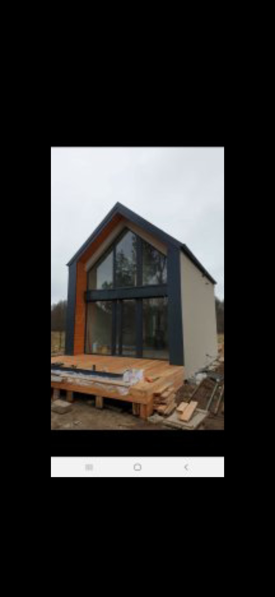 Nowoczesny dom z dużymi oknami i drewnianym tarasem w trakcie budowy. Widoczne elementy konstrukcyjne i materiały budowlane. Minimalistyczny design i naturalne otoczenie.