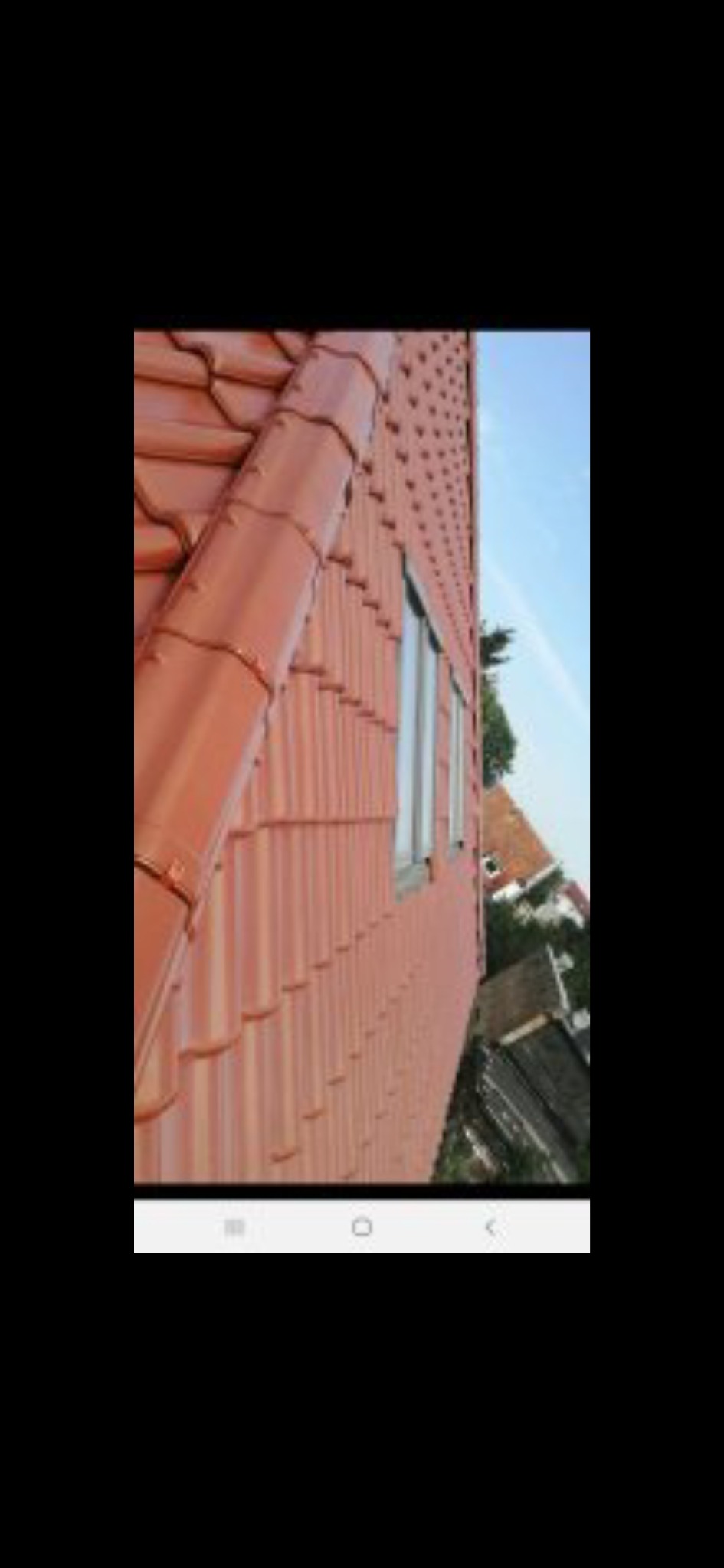 Czerwona dachówka ceramiczna na skośnym dachu domu jednorodzinnego, widok z dołu, z rynnami i oknami. Fragment elewacji i niebo w tle.