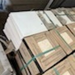 Szybki Błysk - Stos białych płyt i kartonów zabezpieczonych zielonym sznurkiem, ułożonych w magazynie. Widoczne ślady kurzu i zabrudzeń, sugerujące przygotowanie do sprzątania.