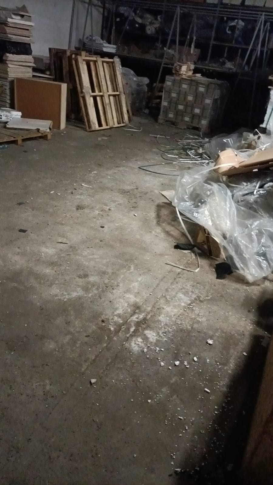 Zabrudzona podłoga w magazynie z paletami, kartonami i folią. Widoczne ślady po remoncie lub opróżnianiu. Słabe oświetlenie, surowy klimat.