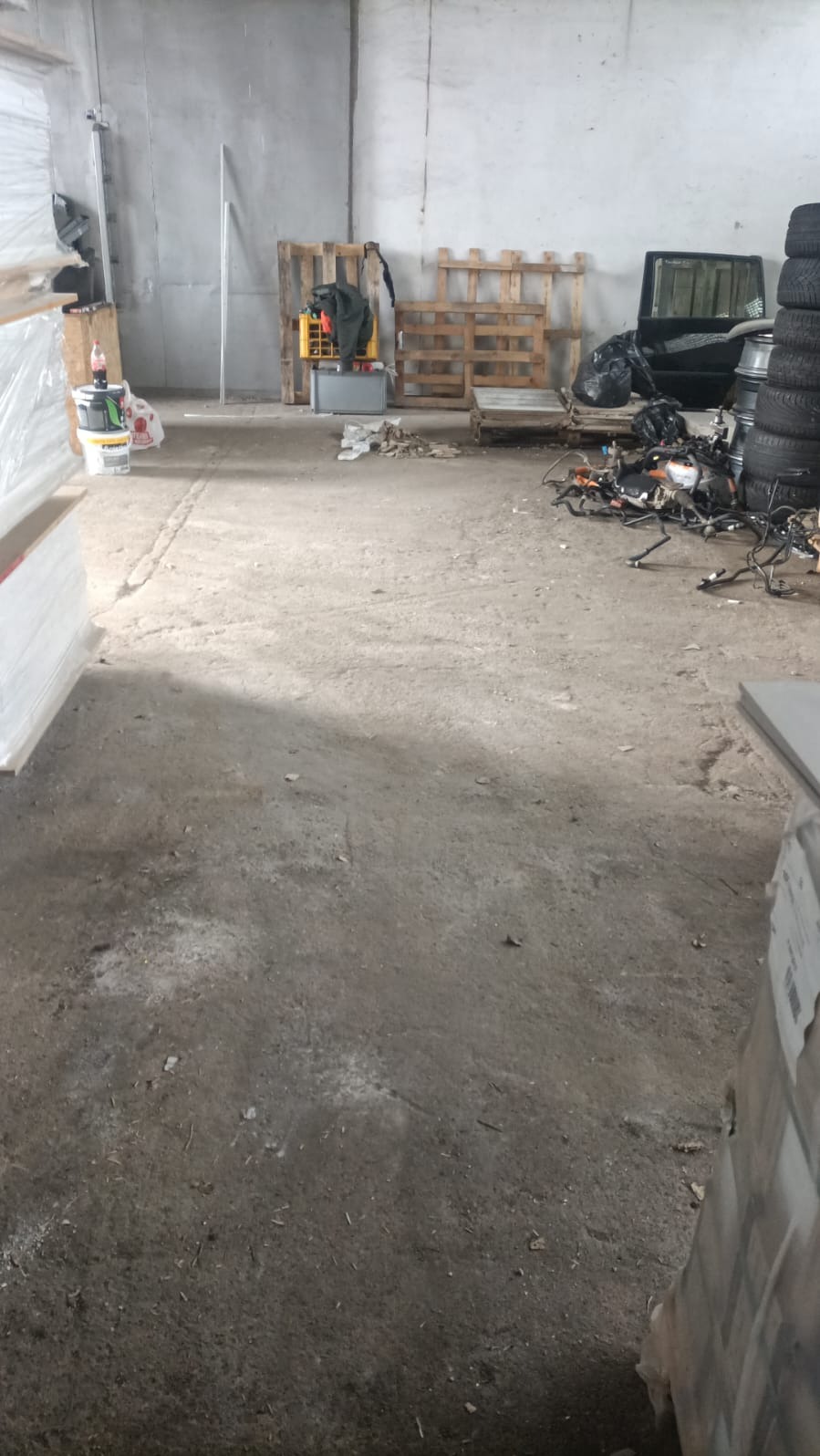 Zabrudzona hala magazynowa z paletami, oponami i elementami samochodowymi. Widoczne ślady użytkowania i pozostałości po pracach. Betonowa podłoga z plamami.