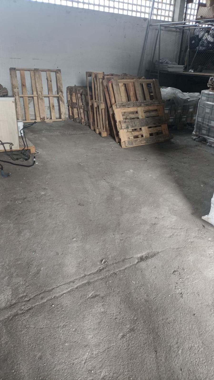 Stos drewnianych palet w magazynie z betonową podłogą i białymi ścianami. Palety ułożone jedna na drugiej, widoczne ślady użytkowania. W tle metalowa konstrukcja.