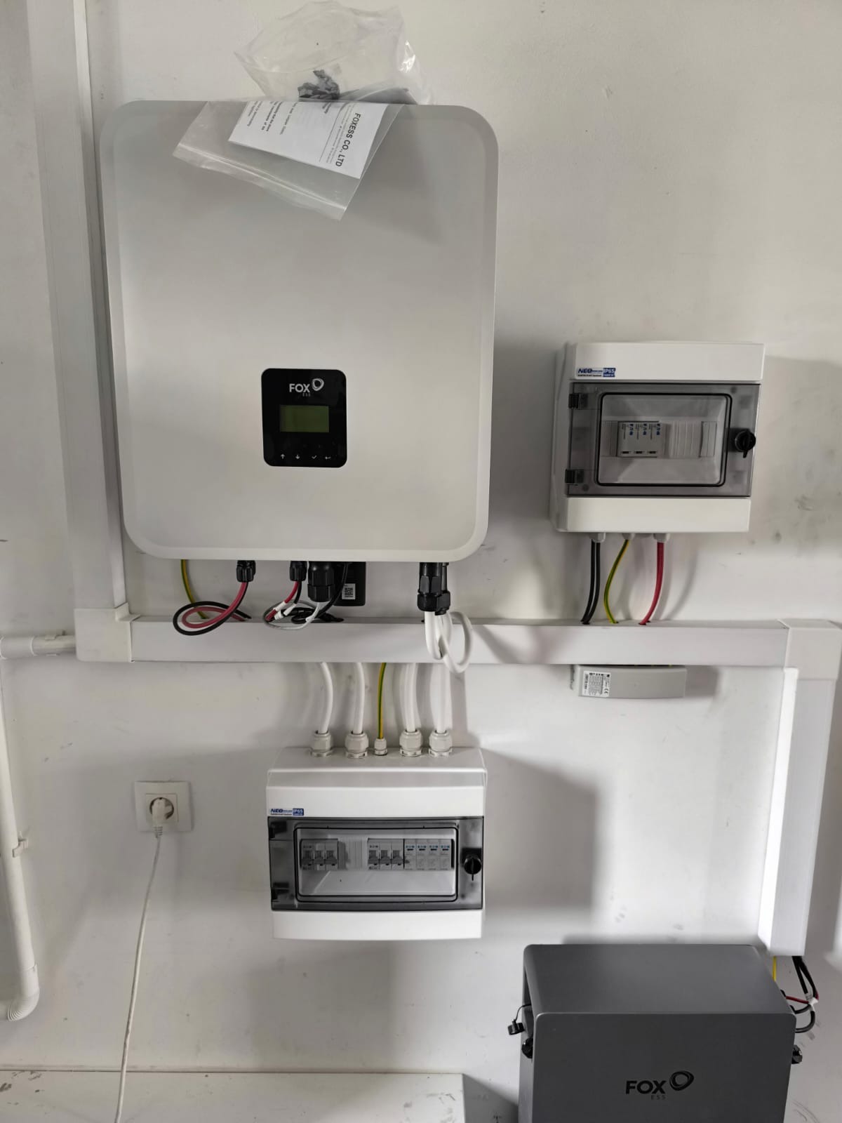 Zamontowany inwerter fotowoltaiczny Fox oraz rozdzielnice elektryczne na białej ścianie. Widoczne przewody i kable w białych listwach.