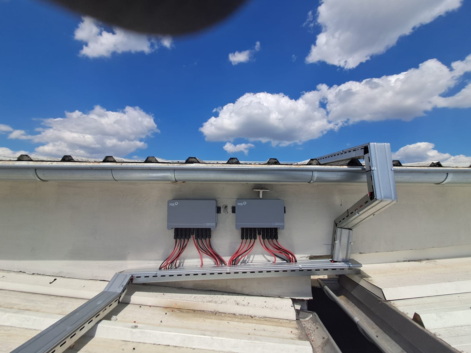 Dwa szare inwertery FOX-D zamontowane na dachu z widocznymi przewodami i elementami montażowymi na tle błękitnego nieba z chmurami.