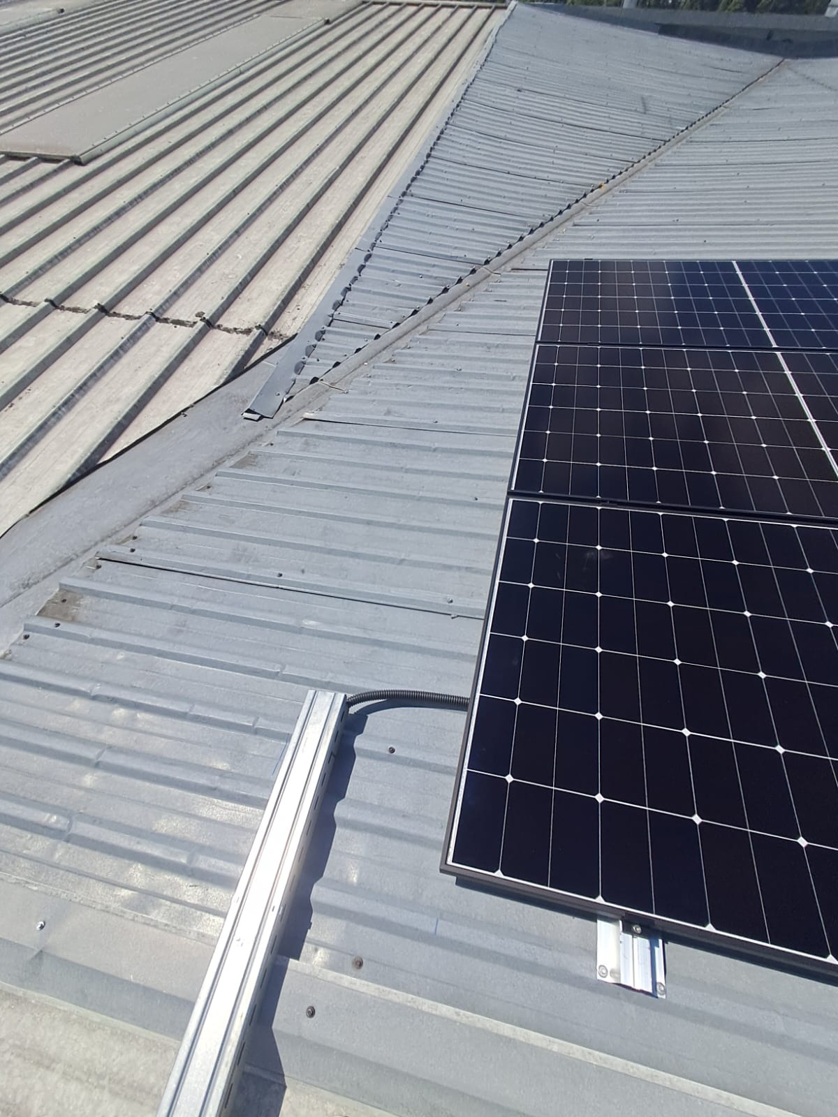 Panele fotowoltaiczne zamontowane na metalowym dachu z widocznymi elementami mocującymi i prowadnicami kablowymi. Ujęcie z góry, oświetlenie dzienne.