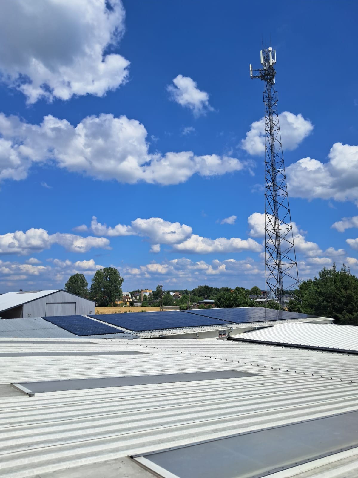 Panele słoneczne na dachu budynku przemysłowego w słoneczny dzień, z wieżą telekomunikacyjną w tle i błękitnym niebem z białymi chmurami.