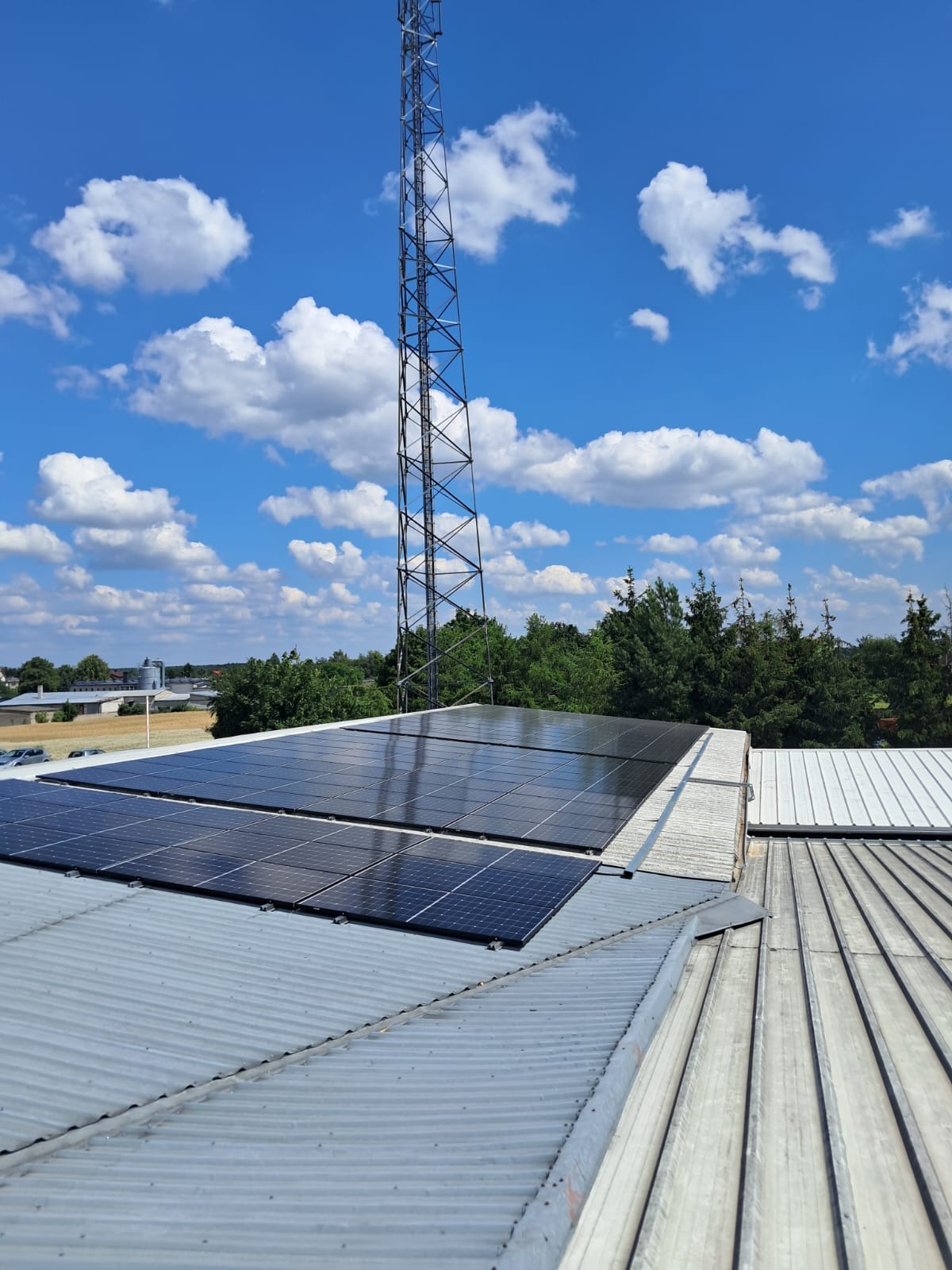 Instalacja fotowoltaiczna na blaszanym dachu budynku, widok z góry, w tle wieża telekomunikacyjna i błękitne niebo z chmurami. Panele w dwóch rzędach.