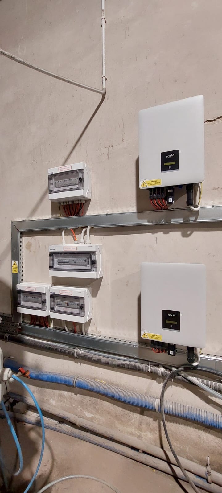 Szara ściana z zamontowanymi skrzynkami elektrycznymi i dwoma białymi falownikami FOXESS. Widoczne kable i rurki instalacyjne. Surowy stan wykończenia wnętrza.
