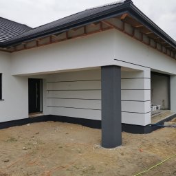 ZAKŁAD OGÓLNOBUDOWLANY Michał Waśkowiak - Nowoczesny dom jednorodzinny z białą elewacją i szarymi akcentami, widok z zewnątrz, w tle budowa. Charakterystyczne poziome pasy na elewacji.