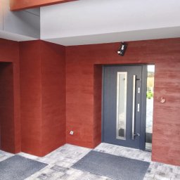 ZAKŁAD OGÓLNOBUDOWLANY Michał Waśkowiak - Nowoczesne wejście do domu z drzwiami w kolorze grafitowym i ścianami wykończonymi w odcieniach czerwieni, imitującymi drewno. Widoczne oświetlenie zewnętrzne.