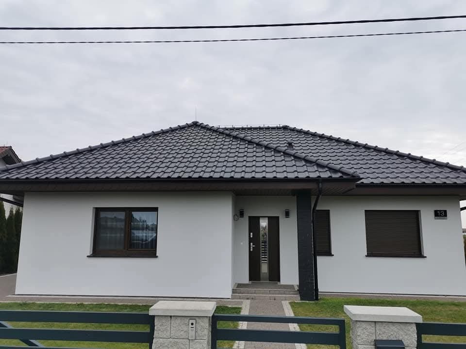 Biały parterowy dom z ciemną dachówką i numerem 13, widoczny z przodu, otoczony zielonym trawnikiem i niskim ogrodzeniem. Nowoczesna architektura.