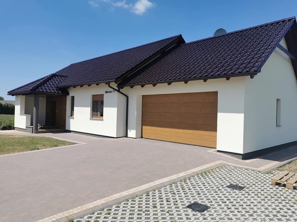 Biały dom jednorodzinny z brązowym dachem i garażem, widok z zewnątrz, kostka brukowa na podjeździe, błękitne niebo w tle. Elewacja po renowacji.