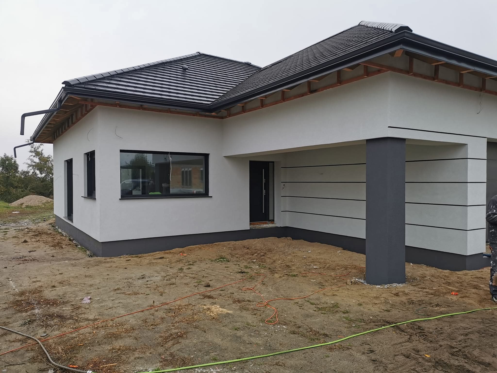 Nowoczesny dom jednorodzinny po tynkowaniu elewacji zewnętrznej w odcieniach bieli i szarości, z widocznymi elementami dachu i otoczeniem w trakcie prac wykończeniowych.