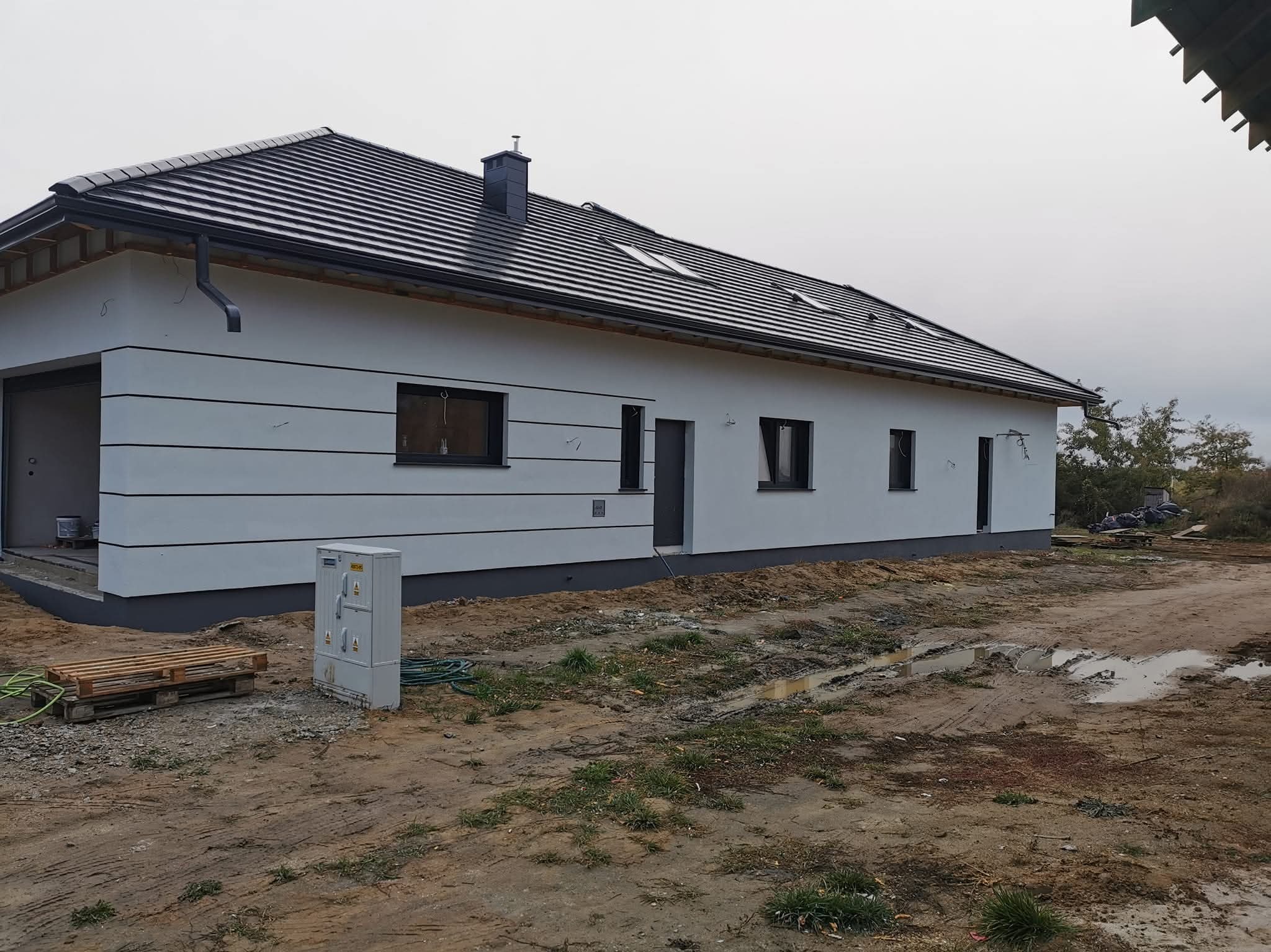 Nowoczesny, parterowy dom z białą elewacją, czarnym dachem i oknami. Widoczne detale: poziome pasy na elewacji, skrzynka elektryczna, paleta i wąż na błotnistym terenie.