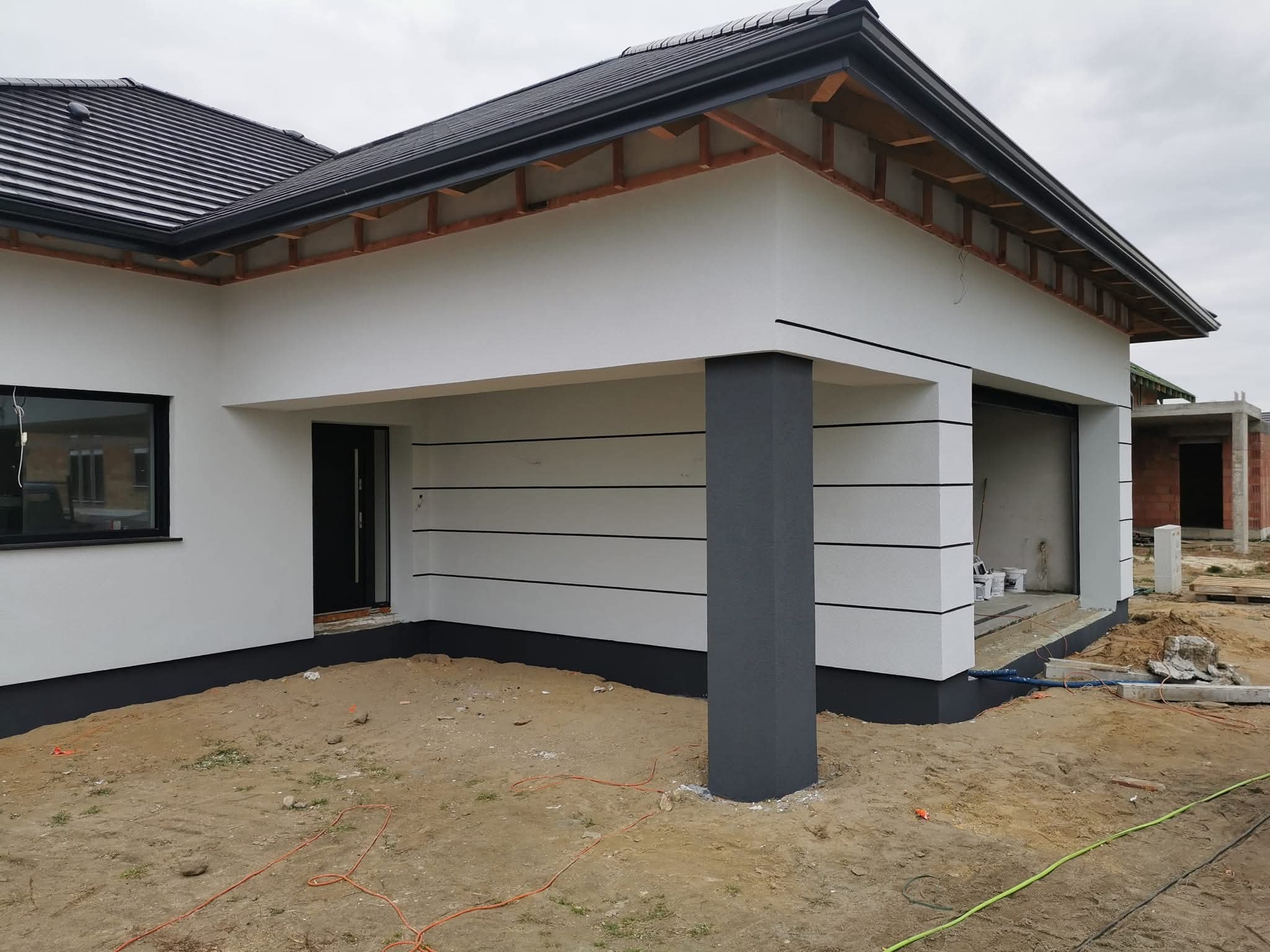 Nowoczesny dom jednorodzinny z białą elewacją i szarymi akcentami, widok z zewnątrz, w tle budowa. Charakterystyczne poziome pasy na elewacji.