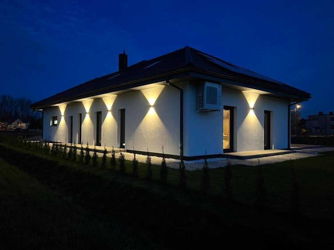Oświetlona elewacja domu jednorodzinnego nocą. Biały tynk, czarne ramy okienne, symetryczne rozmieszczenie lamp elewacyjnych. Widoczny klimatyzator na ścianie.