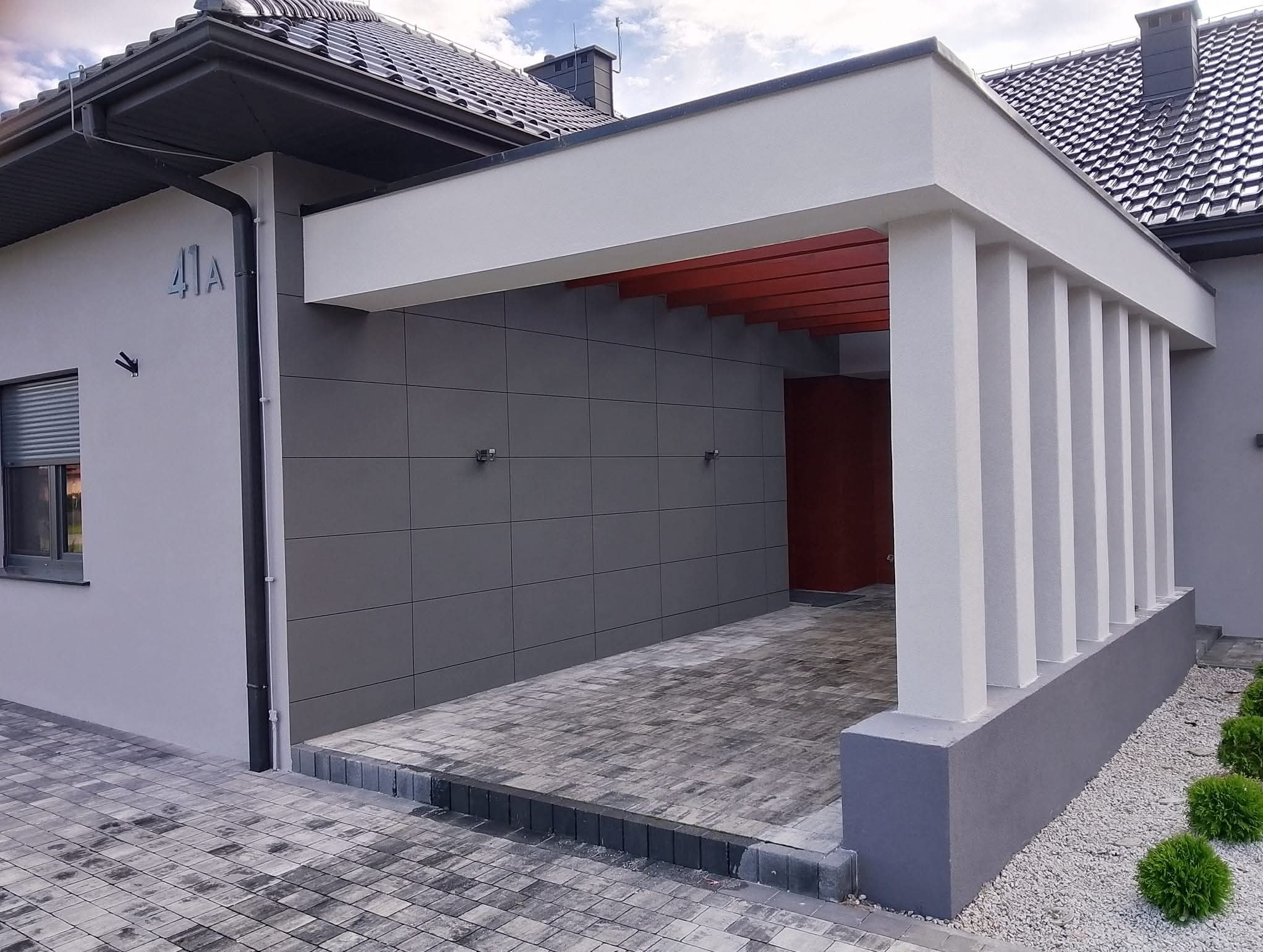 Nowoczesna elewacja domu z podjazdem w Głuchowie. Szare płytki elewacyjne, białe kolumny i elementy konstrukcyjne. Minimalistyczny design i staranne wykonanie.