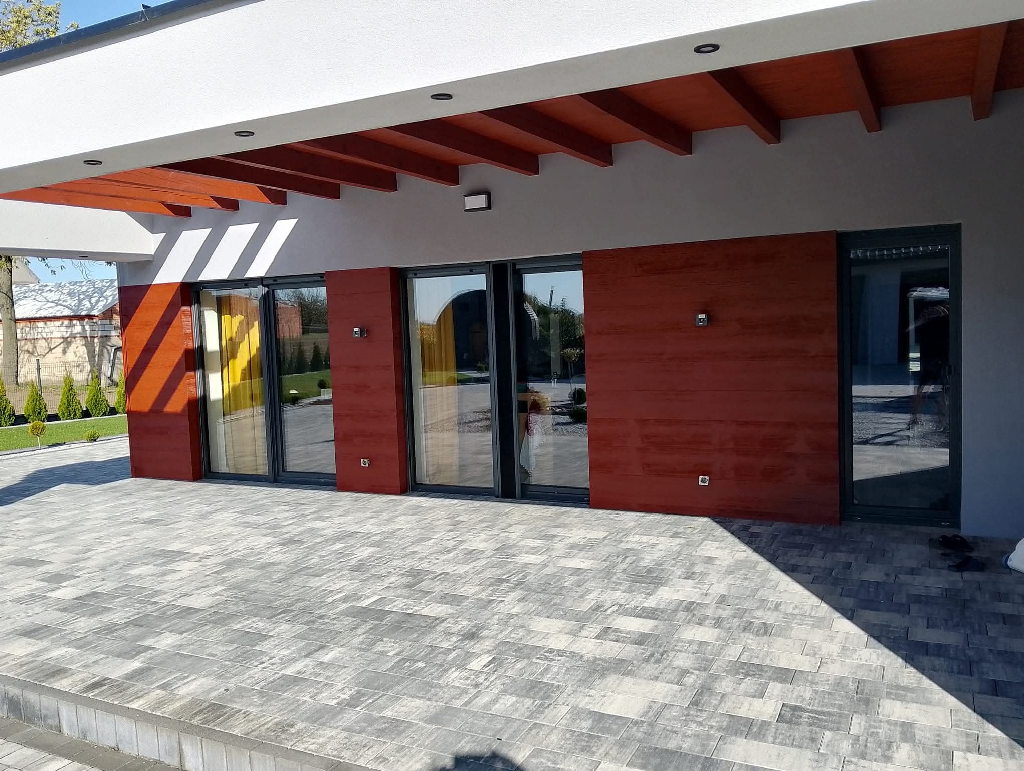 Nowoczesny dom z elewacją w odcieniach szarości i czerwonego drewna. Widoczne duże okna i taras wyłożony kostką brukową. Elementy drewniane pod dachem.