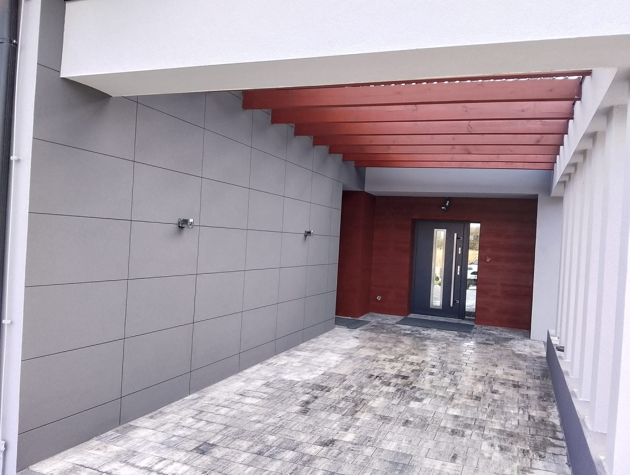 Nowoczesna elewacja domu z szarymi płytami i drewnianą pergolą nad wejściem. Widoczna grafitowa stolarka drzwiowa i szara kostka brukowa.