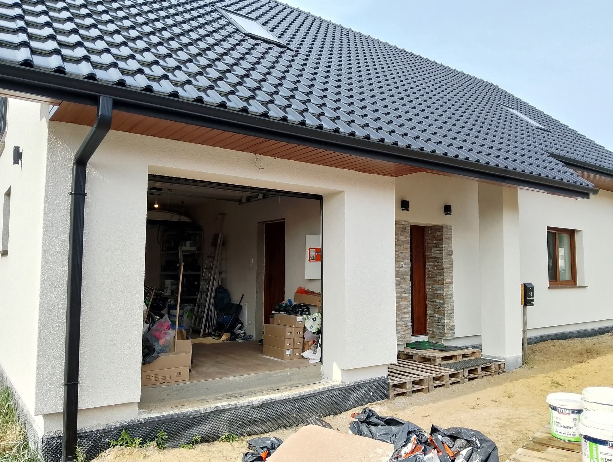 Jasny dom jednorodzinny z ciemną dachówką i wykończoną elewacją. Widoczne palety, worki i puszki z tynkiem silikonowym. Otwarty garaż z narzędziami w środku.