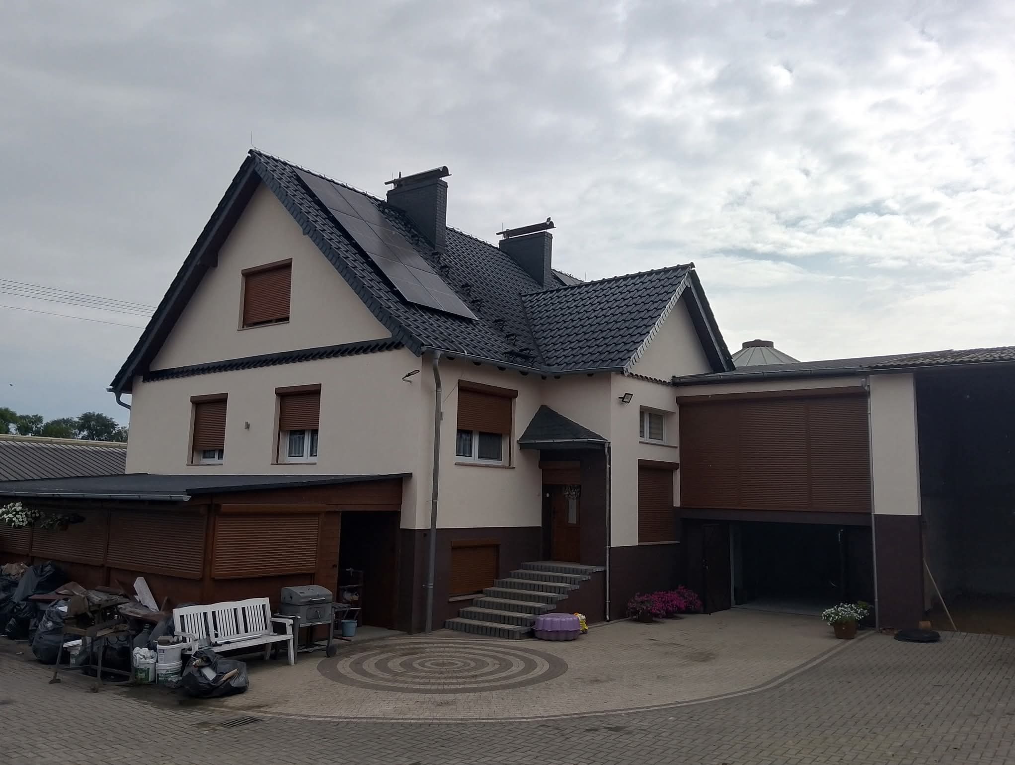 Dwukondygnacyjny dom z panelami słonecznymi na dachu i brązowymi roletami. Widoczne schody wejściowe i garaż z otwartą bramą. Na podwórku biała ławka i narzędzia.