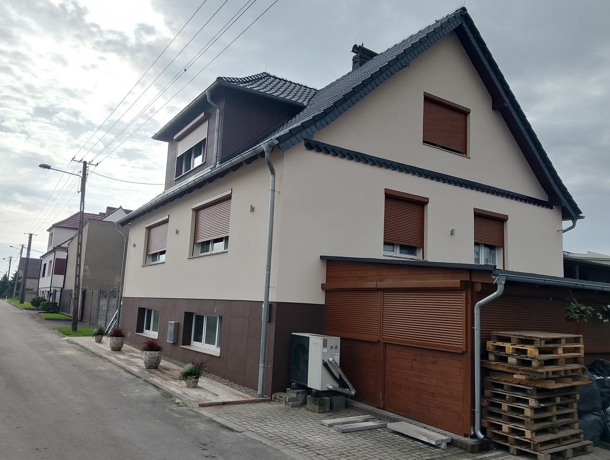 Dwukondygnacyjny dom z beżową elewacją i brązowymi akcentami, w tym rolety okienne. Widoczne palety i klimatyzator na zewnątrz. Pochmurne niebo w tle.