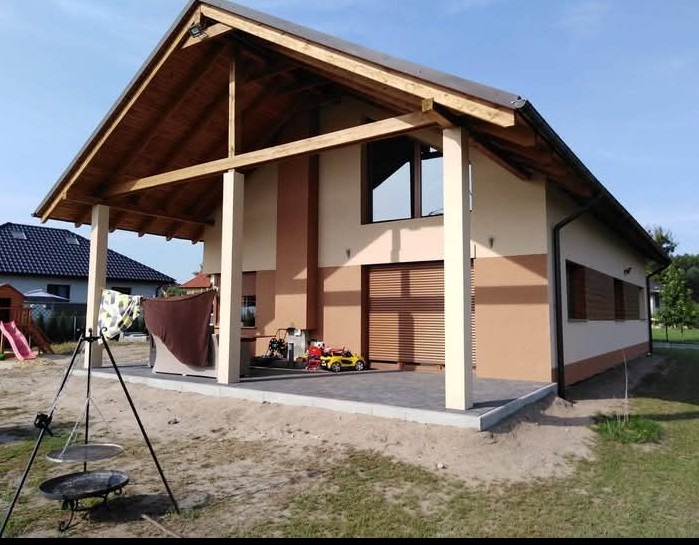 Dwukolorowa elewacja domu z drewnianą konstrukcją zadaszenia tarasu, widok z zewnątrz, w tle plac zabaw i zieleń.