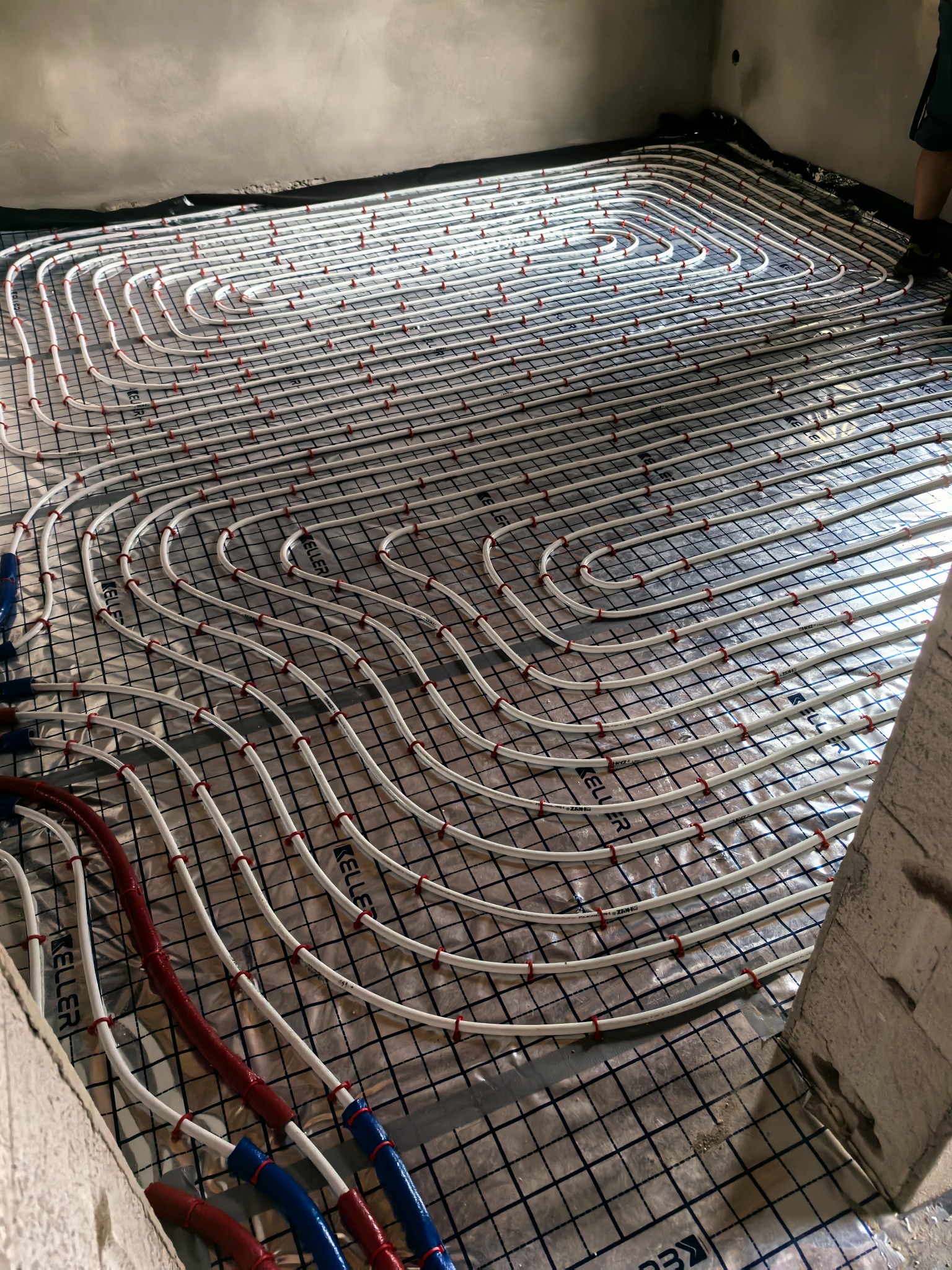 Białe rury ogrzewania podłogowego, równolegle ułożone na folii z siatką, przymocowane czerwonymi uchwytami. Fragment ściany w prawym dolnym rogu.