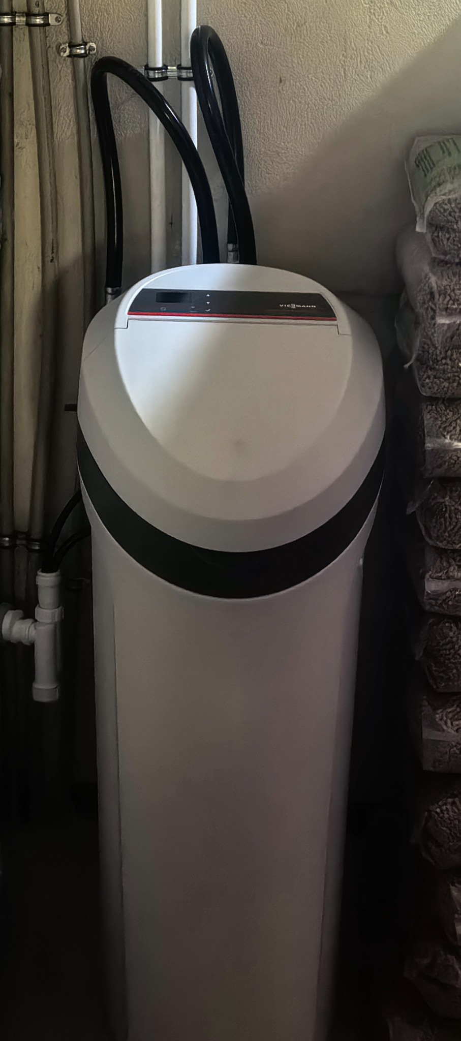 Biały zmiękczacz wody Viessmann z czarnymi rurami w kotłowni, obok worki z pelletem. Widoczne elementy instalacji hydraulicznej.