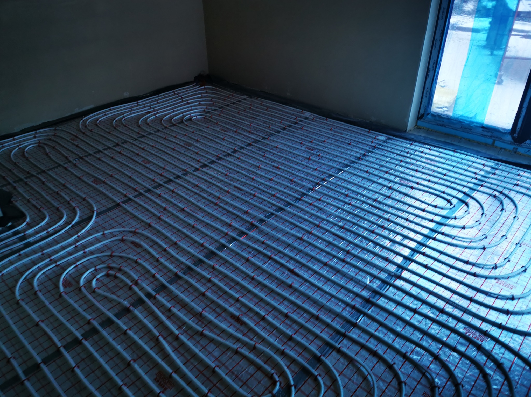 Białe rury ogrzewania podłogowego ułożone na folii w pomieszczeniu w Międzychodzie. Widok z góry, rury tworzą regularny wzór, przygotowane do zalania posadzką.