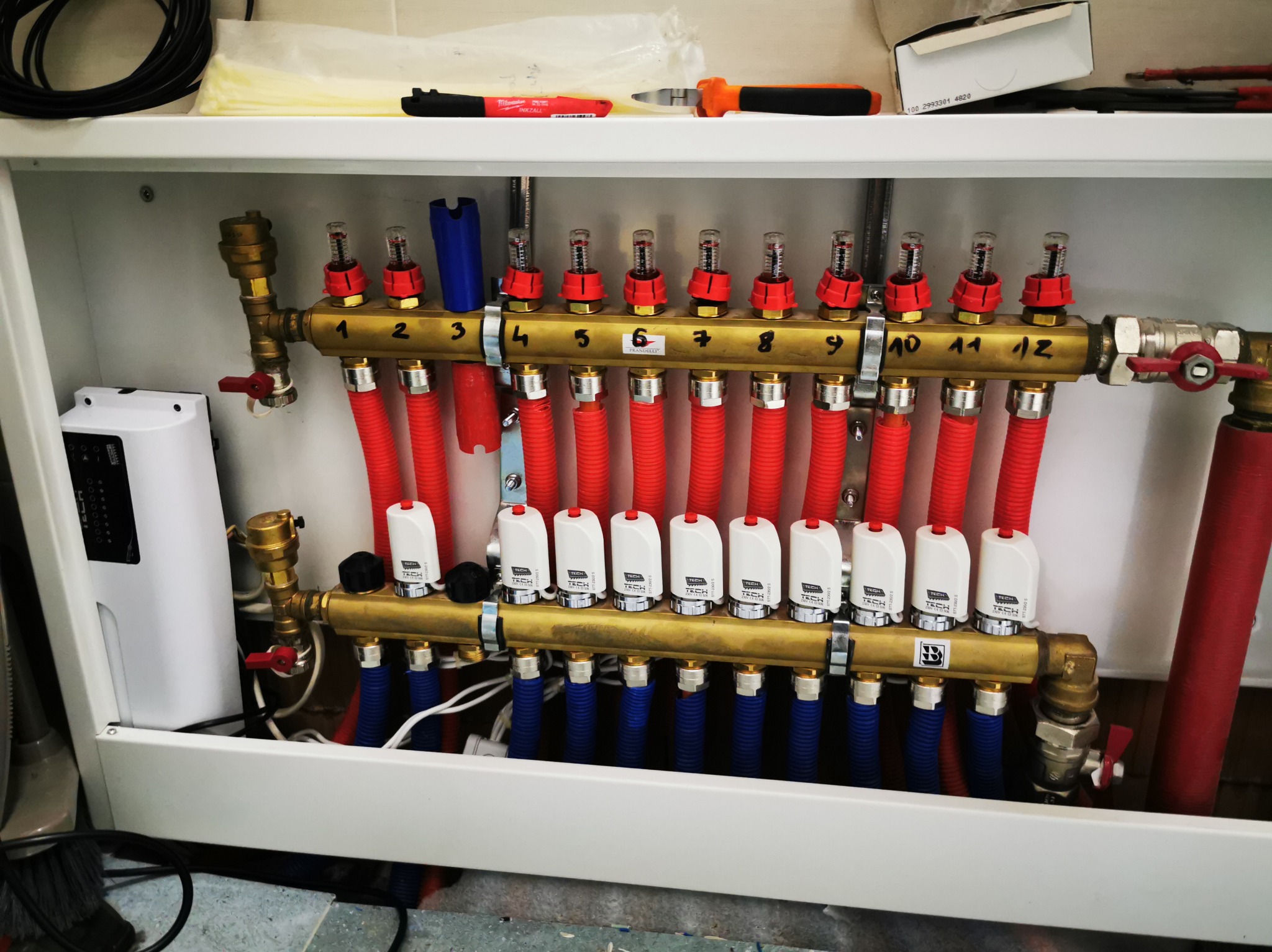 Rozdzielacz ogrzewania podłogowego z zaworami termostatycznymi i rurami w kolorze czerwonym i niebieskim, umieszczony w białej szafce instalacyjnej.