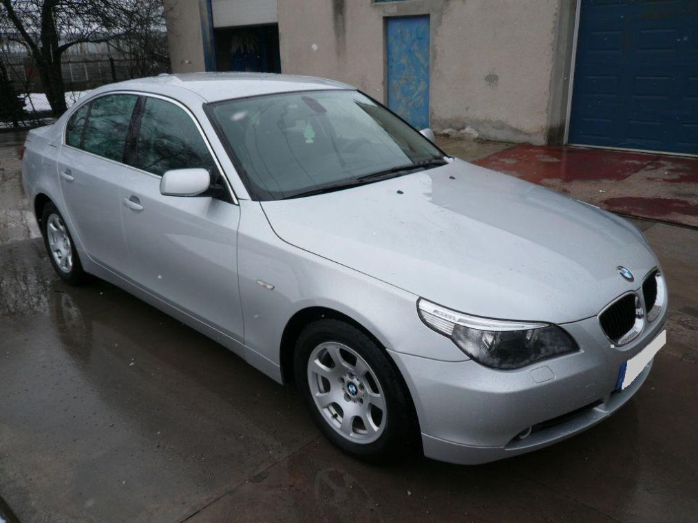 Srebrny sedan BMW serii 5 zaparkowany przed budynkiem z niebieskimi drzwiami garażowymi i drzewem bez liści w tle.