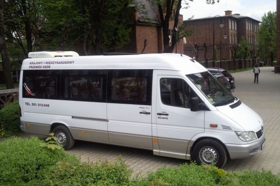 Biały bus Mercedes Sprinter z nadrukiem 'Krajowy i Międzynarodowy Przewóz Osób' oraz numerem telefonu, zaparkowany na tle ceglanego budynku i zieleni.