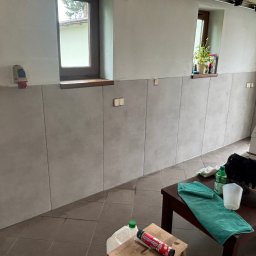 Adrian kokot - Ściana wyłożona szarymi płytami w pomieszczeniu z oknami. Widoczne narzędzia i akcesoria na stole. Płytki ułożone do pewnej wysokości, powyżej ściana pomalowana na biało.