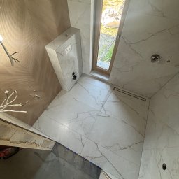 Adrian kokot - Wykończona łazienka z białymi płytkami imitującymi marmur, widok z góry na podłogę z odpływem liniowym i fragment ściany z drewnopodobną fakturą.