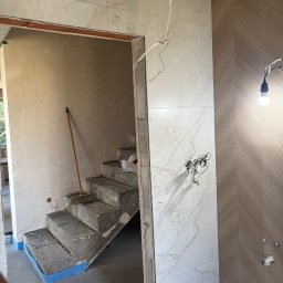 Adrian kokot - Wnętrze w trakcie prac wykończeniowych: marmurowe płytki na ścianie, surowe schody z niebieską taśmą zabezpieczającą, widoczne przewody elektryczne i włącznik światła.