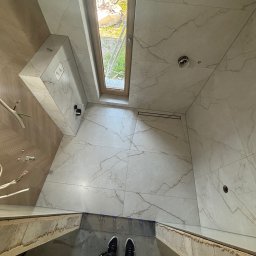 Adrian kokot - Wykończona łazienka z marmurowymi płytkami, widok z góry na podłogę i fragment osoby. Uwagę przykuwa okno dachowe i minimalistyczny odpływ liniowy.