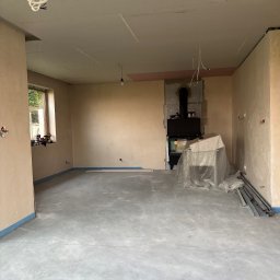 Adrian kokot - Wnętrze w trakcie prac wykończeniowych: surowe ściany, wylewka podłogowa, kominek zabezpieczony folią. Widoczne przewody elektryczne i materiały budowlane.