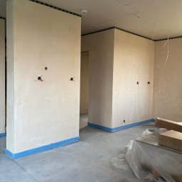 Adrian kokot - Wnętrze w trakcie prac wykończeniowych: ściany po szpachlowaniu, zabezpieczone taśmą malarską, widoczne puszki elektryczne, sufit z płyt karton-gips.