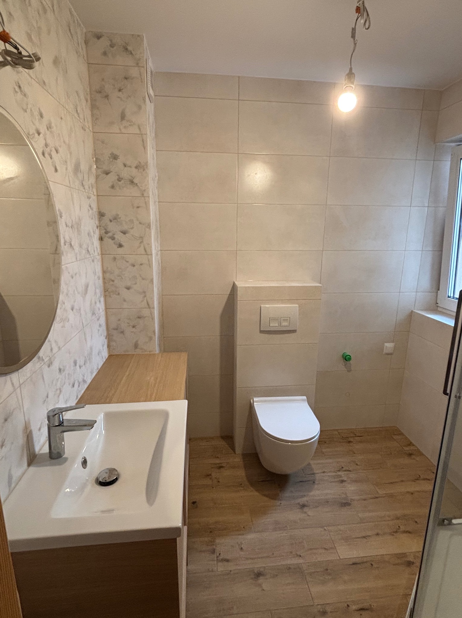 Jasna łazienka po remoncie: beżowe płytki ścienne, drewnopodobna podłoga, biała umywalka z szafką i toaleta podtynkowa. Minimalistyczny design, ciepłe oświetlenie.