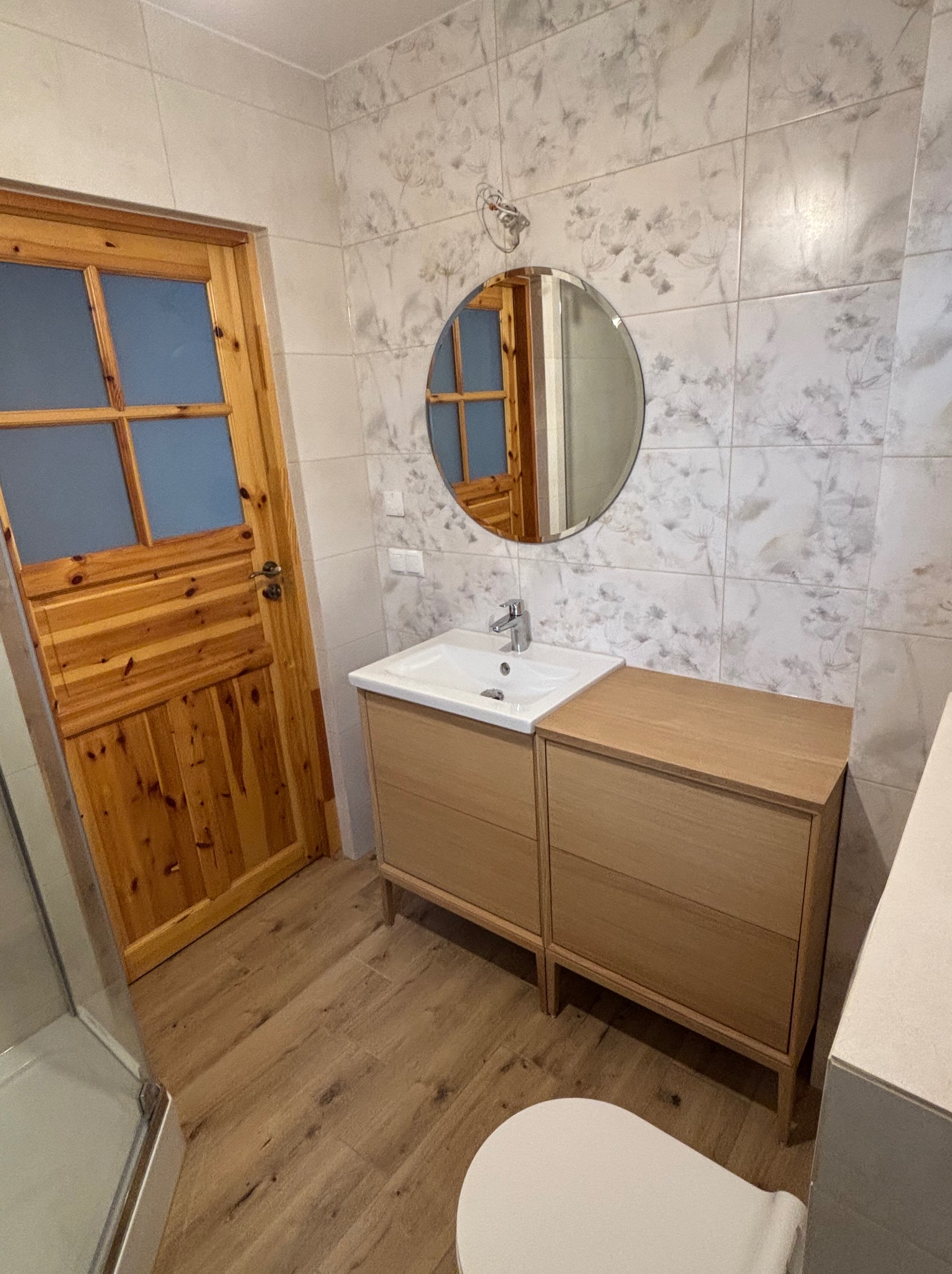 Jasna łazienka w Pszczynie z drewnianymi akcentami: drzwi z szybkami, szafka pod umywalką i podłoga imitująca drewno. Na ścianach płytki z motywem roślinnym.