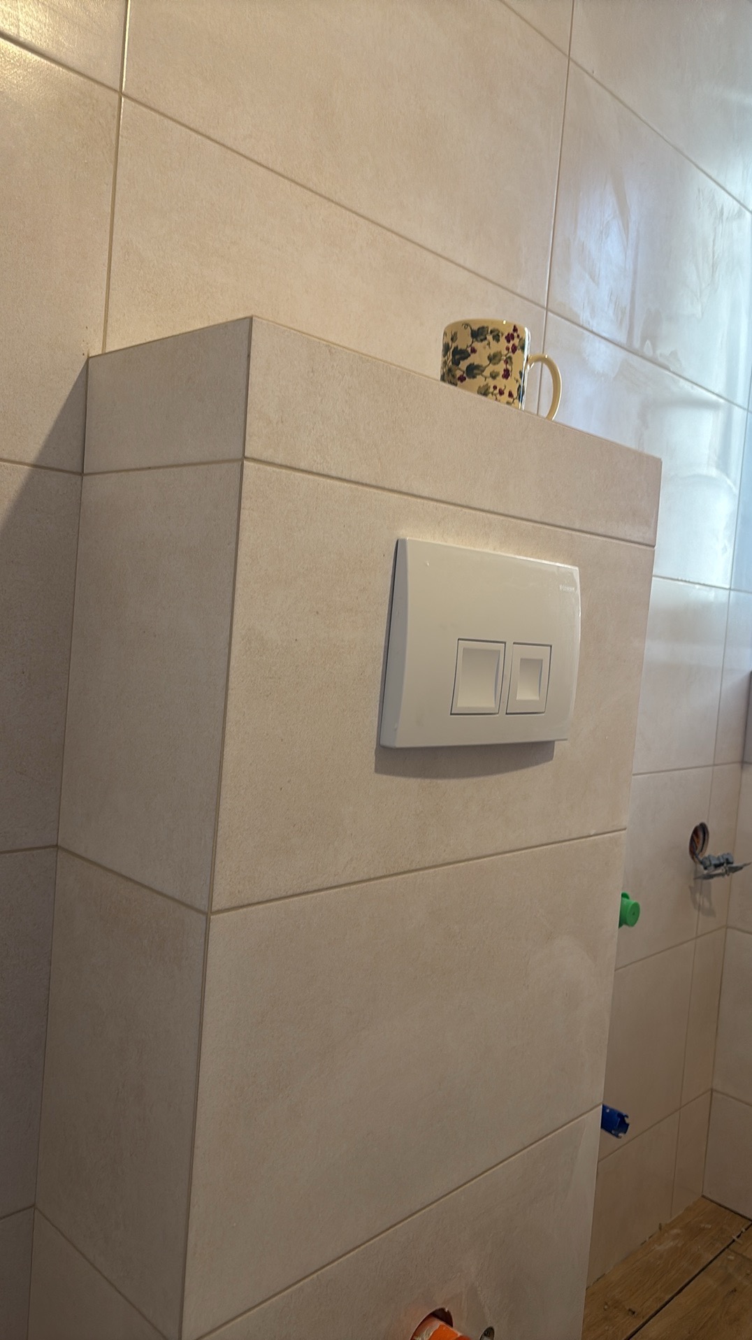 Obudowa WC z beżowych płytek, widoczny przycisk spłukujący i kubek na górze. Ujęcie z bliska, prezentujące detale wykończenia w łazience.