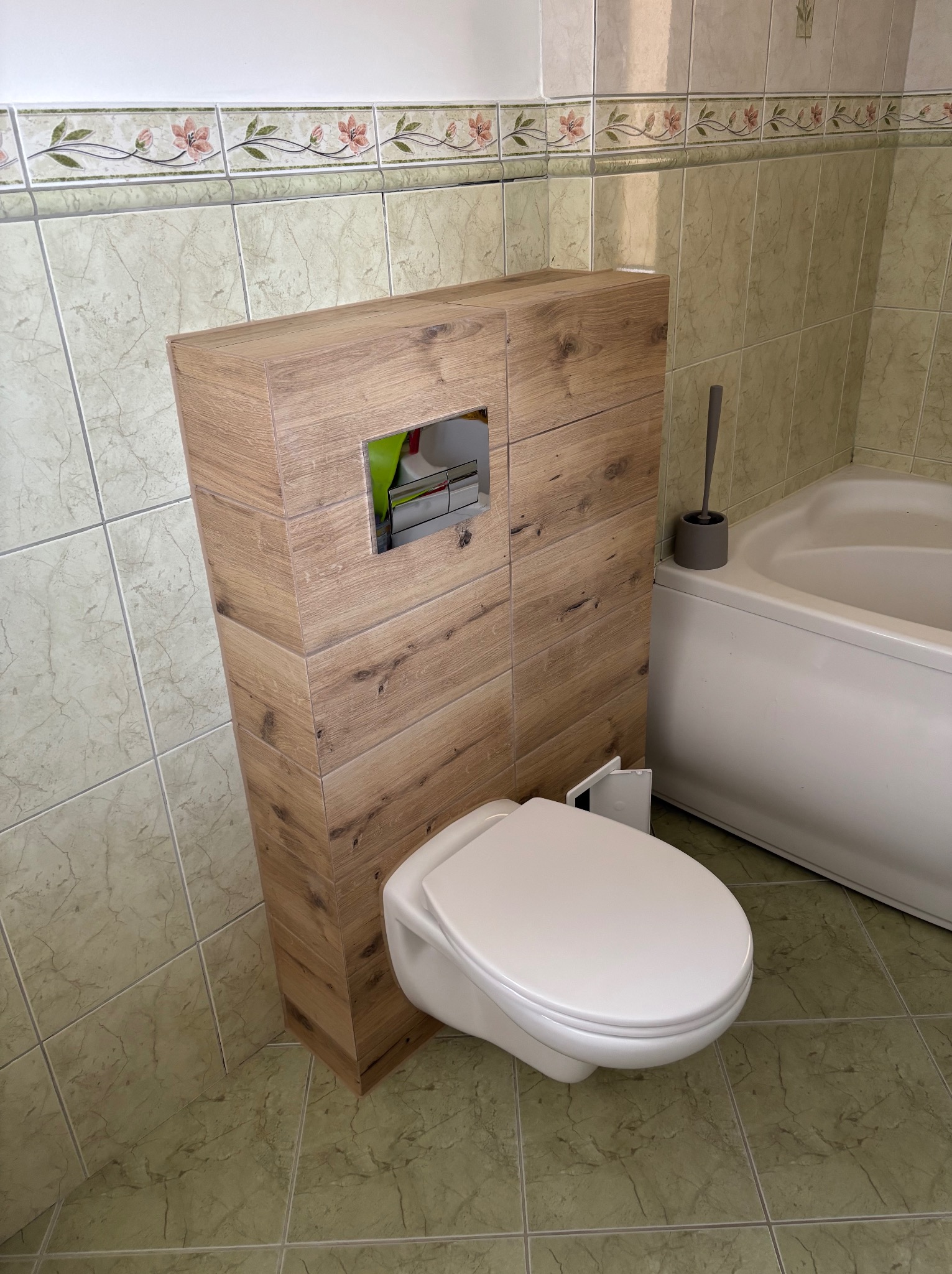 Nowoczesna toaleta podwieszana z zabudową z płytek imitujących drewno, w tle wanna i płytki ścienne z delikatnym wzorem kwiatowym. Ukończona aranżacja łazienki.