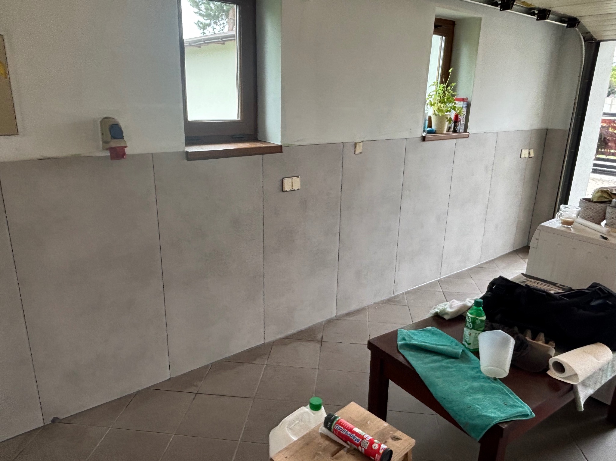 Ściana wyłożona szarymi płytami w pomieszczeniu z oknami. Widoczne narzędzia i akcesoria na stole. Płytki ułożone do pewnej wysokości, powyżej ściana pomalowana na biało.